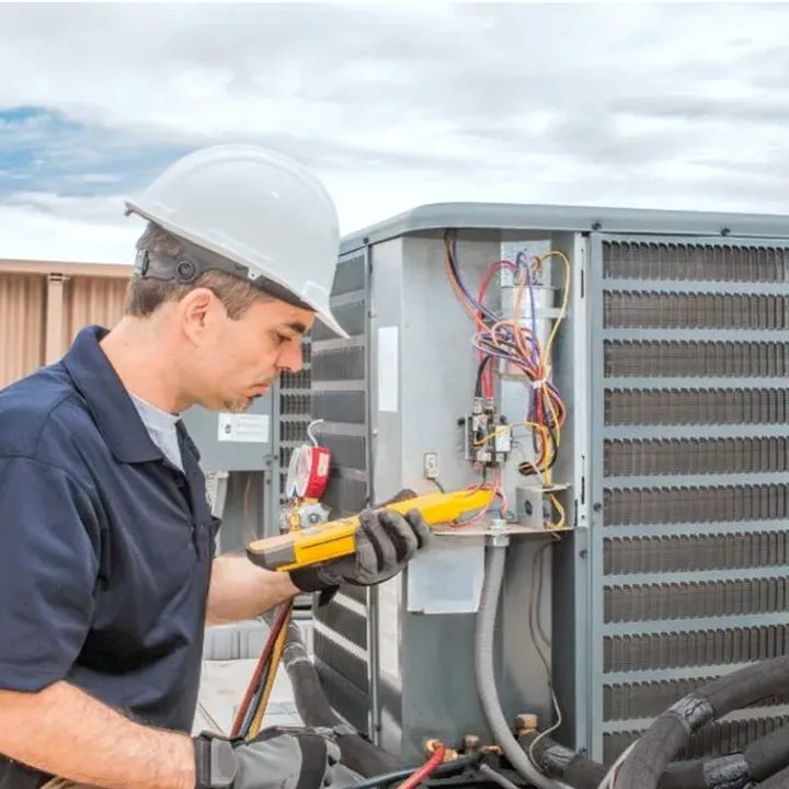 HVAC technician in a hard hat testing an outdoor air conditioner with a multimeter